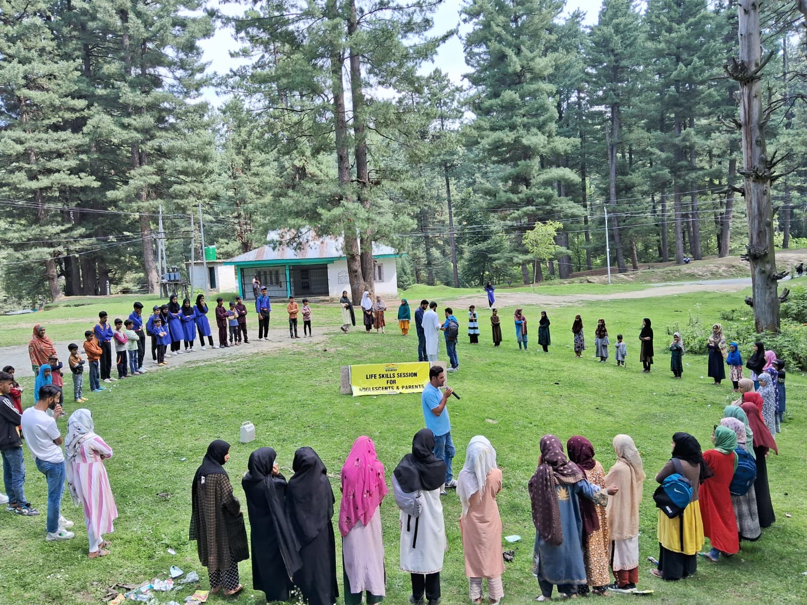 A facilitator addressing a large circle of adolescents and parents during a KOSHISH life skills session in a pine forest