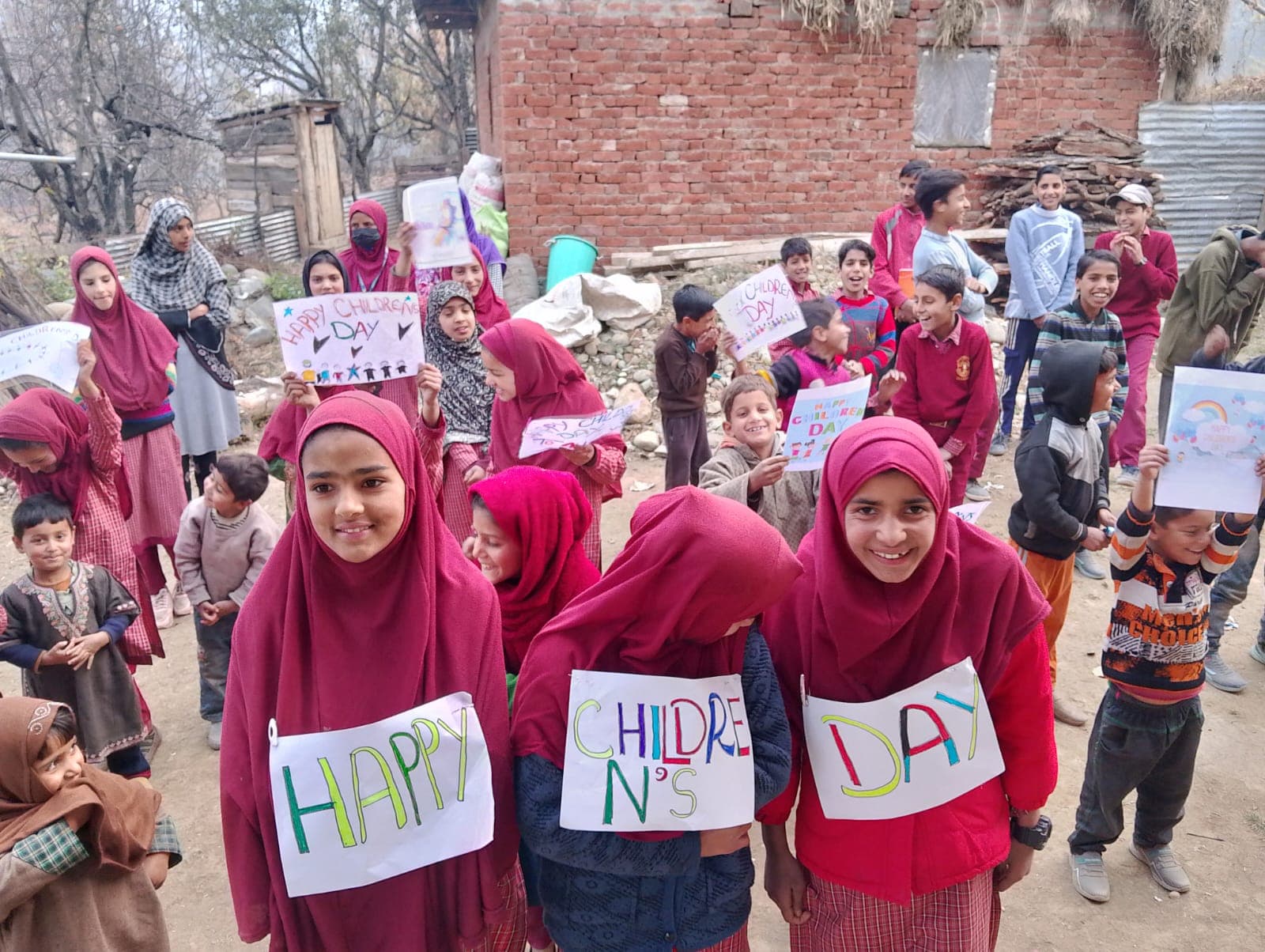 Girls in maroon headscarves holding hand-lettered Happy Children's Day signs in a village street