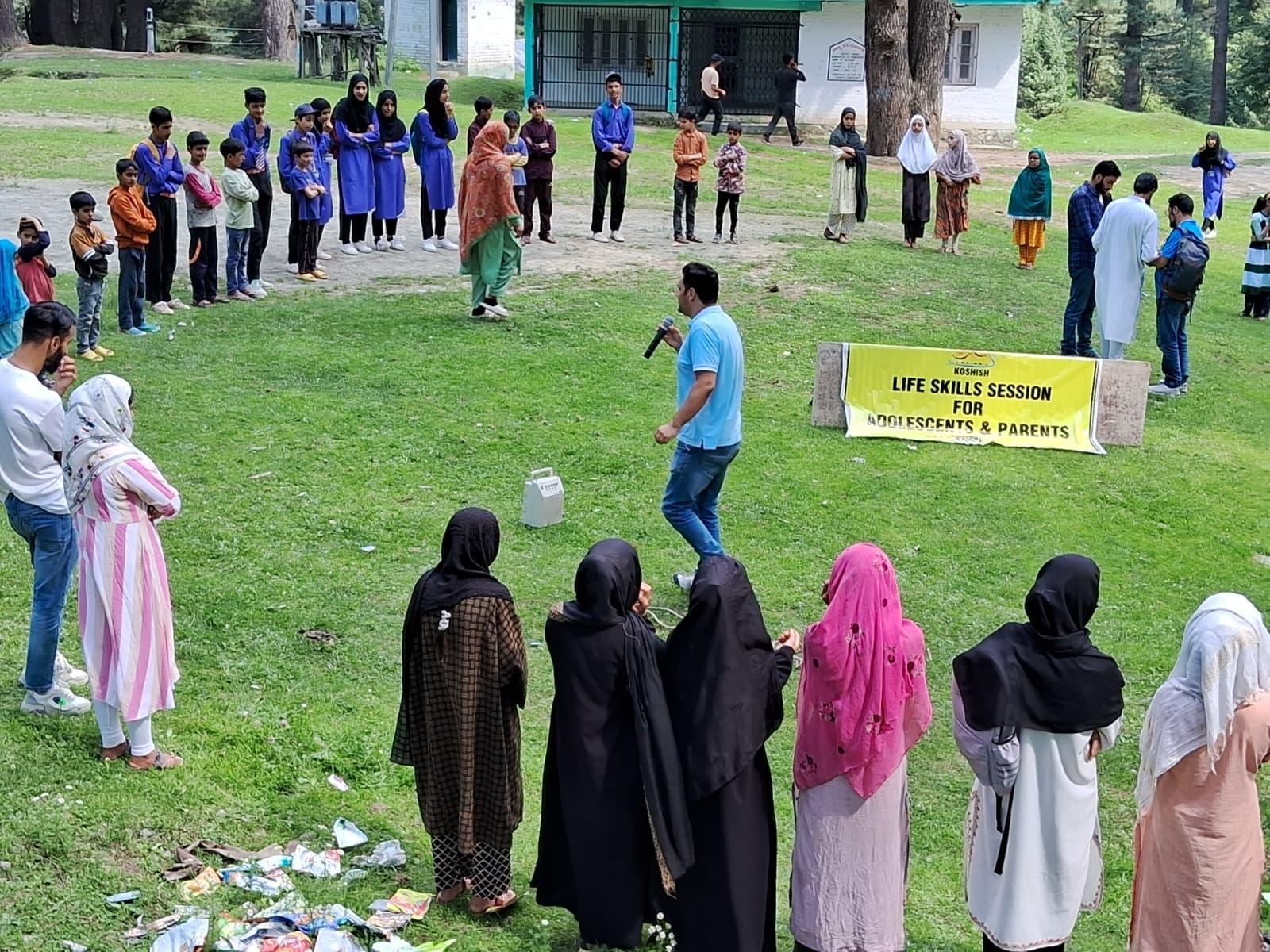 A wide view of adolescents and parents forming a circle on a grass field during a KOSHISH life skills session