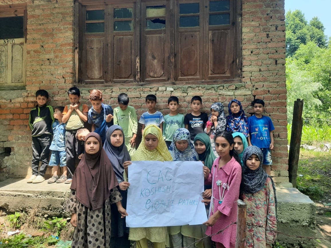 Children holding a CAC KOSHISH Gogji Pathri sign in front of a brick building