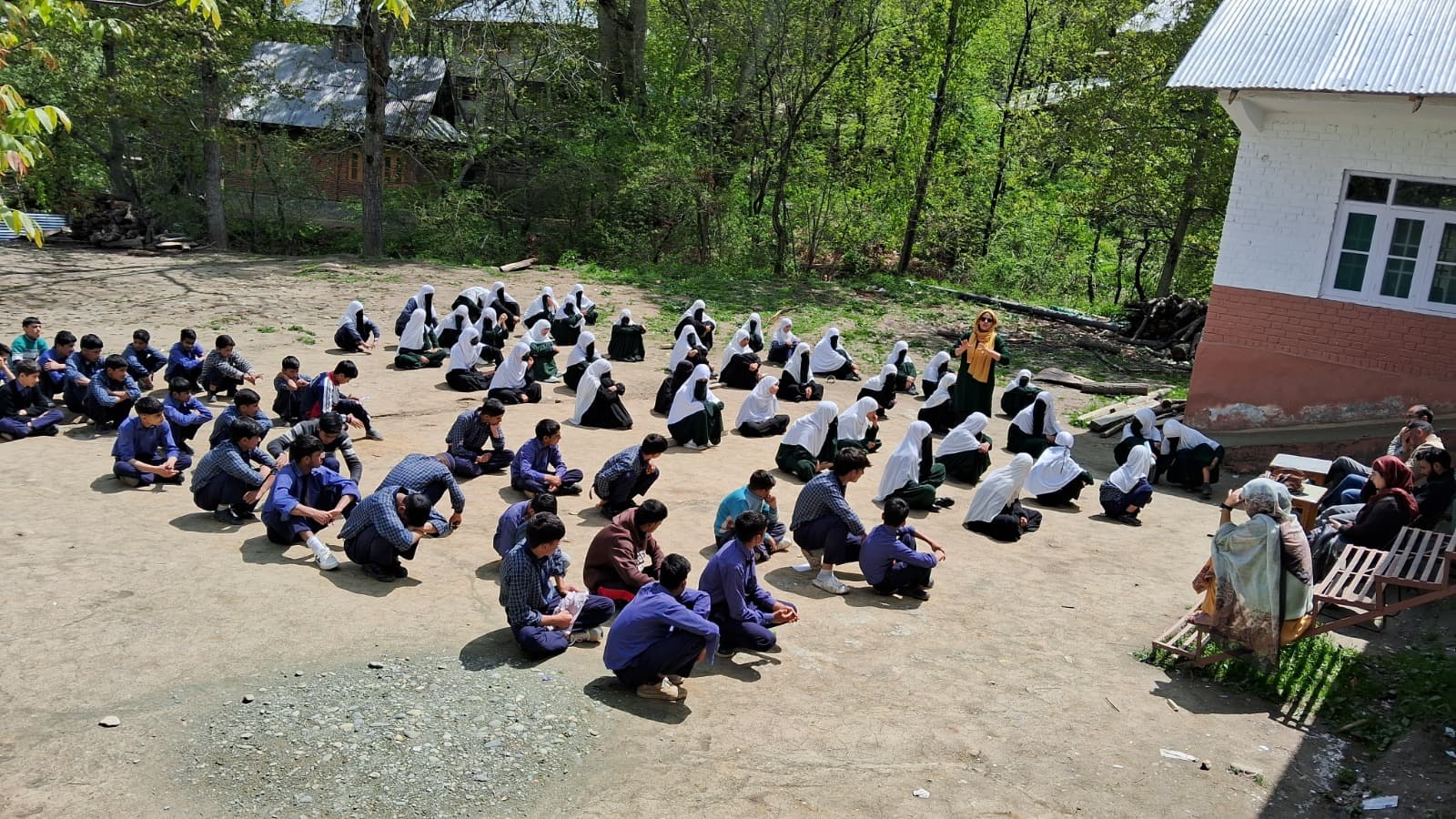 Students seated in rows in a school courtyard during a KOSHISH awareness session led by a facilitator