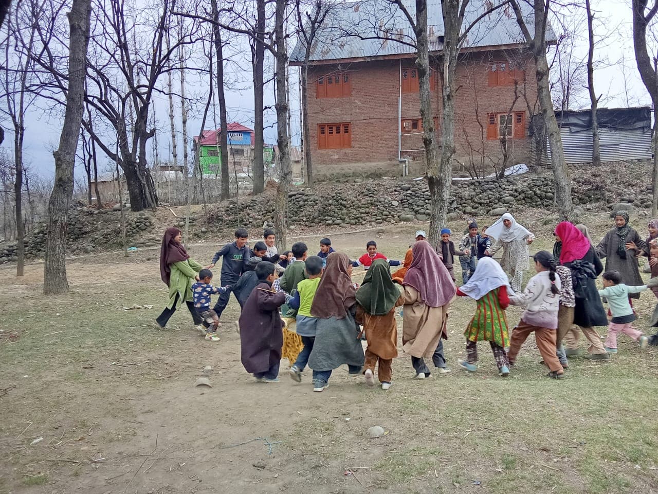 A facilitator addressing students seated at desks on a school lawn in a hillside village