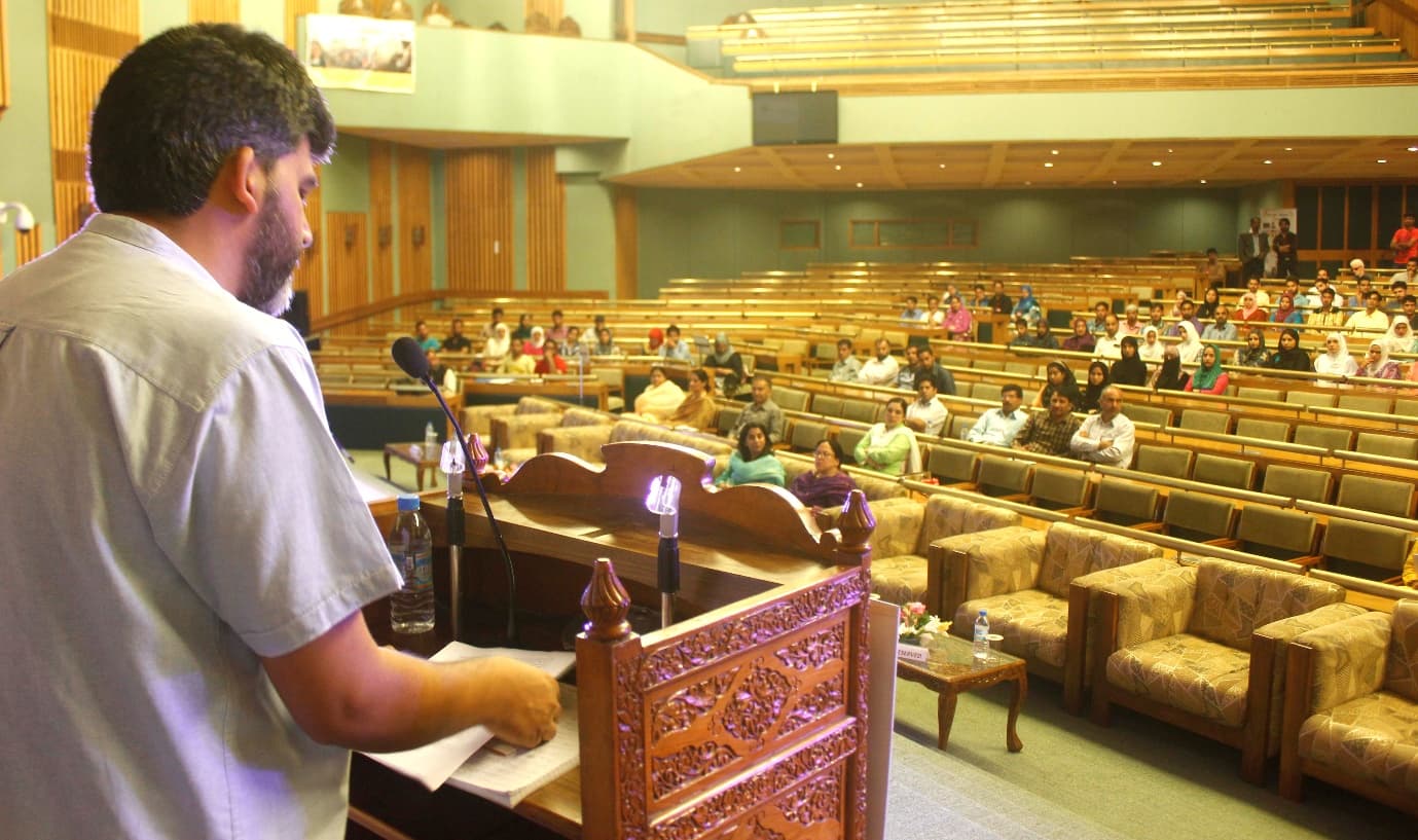 A speaker addressing a full auditorium from a wooden lectern
